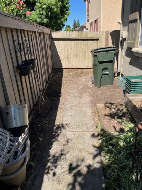 Narrow concrete path between wooden fences, trash can, and house with plants.