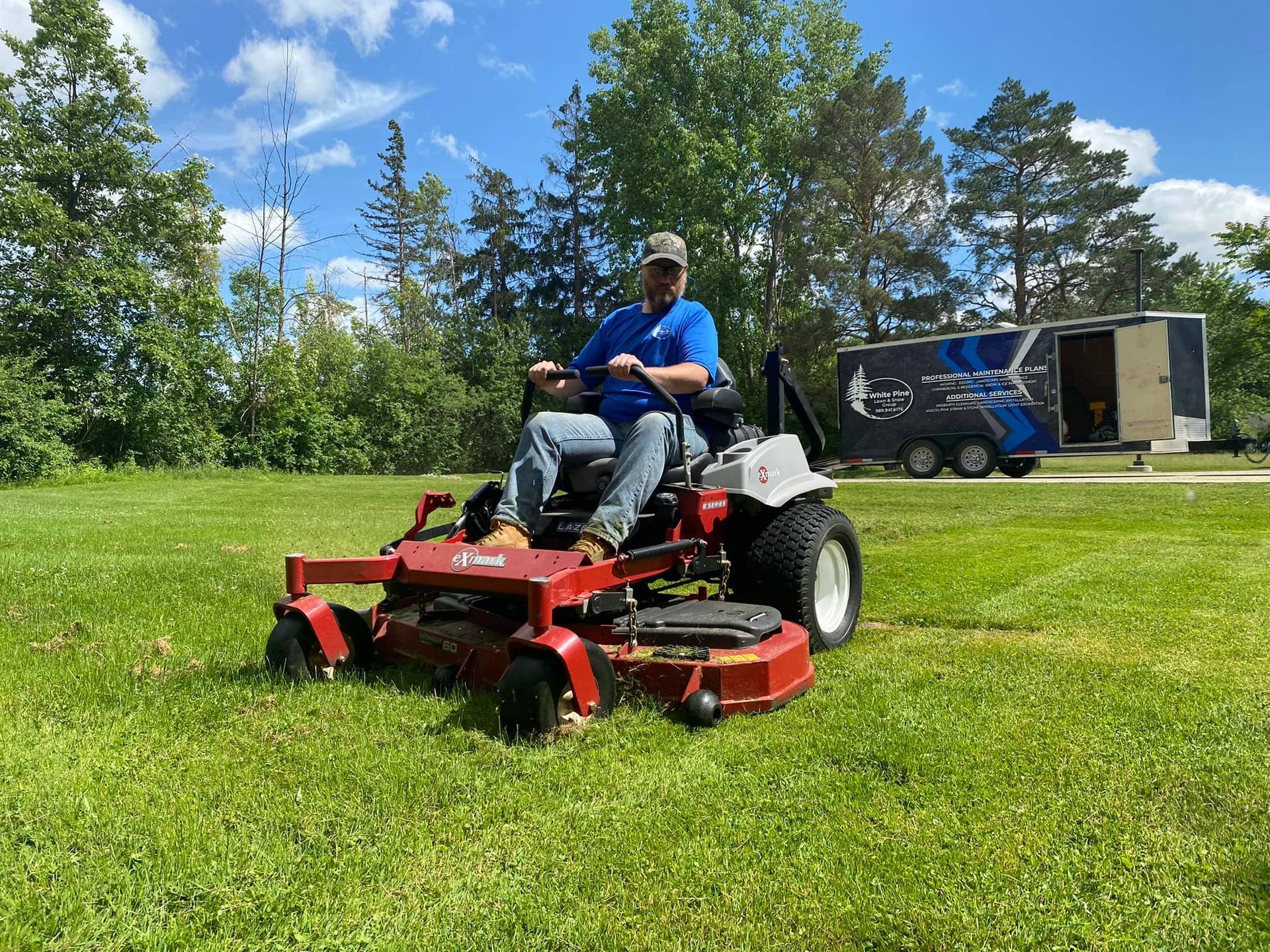 Man on red riding mower cutting grass in a sunny field near trees and a trailer.