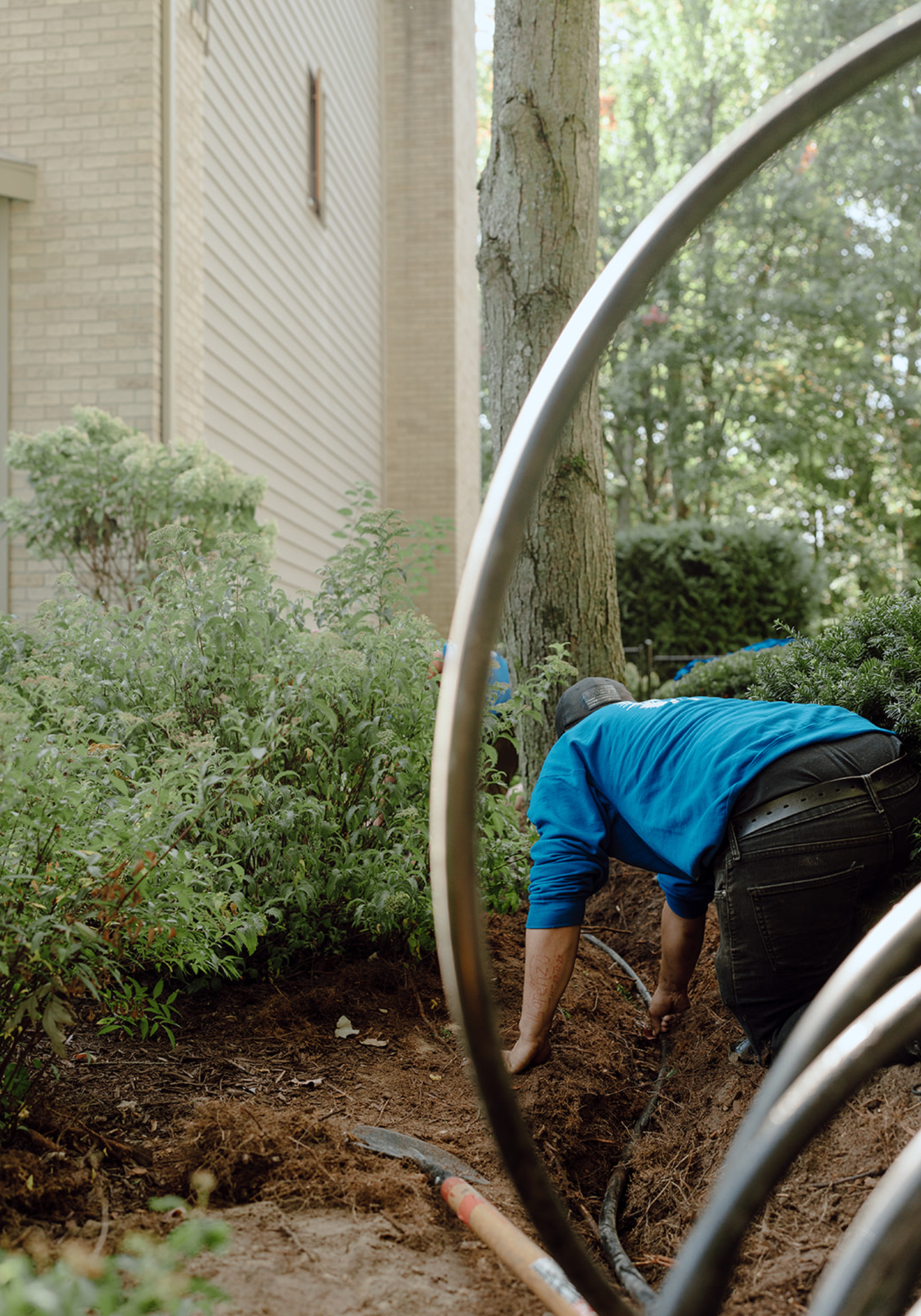 Person in blue jacket bending over in a garden beside a building and metal hoops