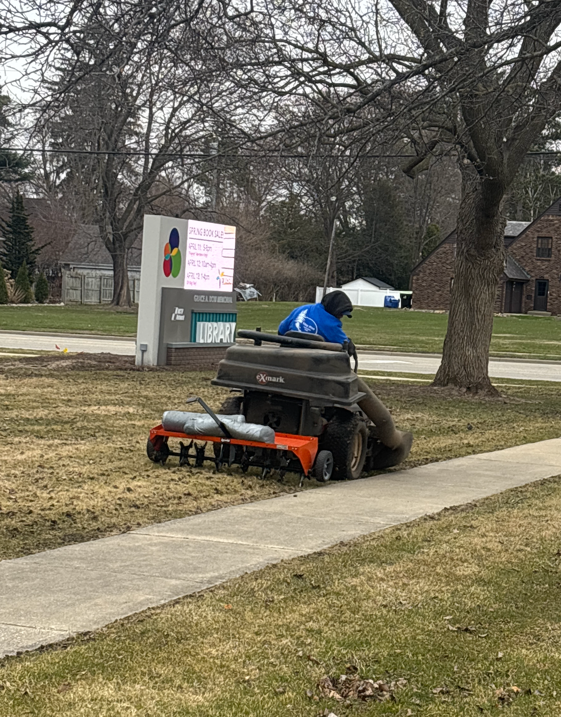 Orange snowblower clearing a sidewalk beside a grassy path in a park-like area