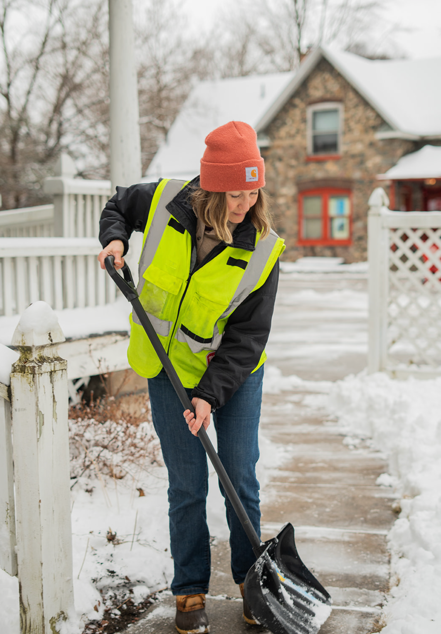 Person shoveling snow on a snowy sidewalk beside a white fence, wearing a neon jacket and orange beanie.