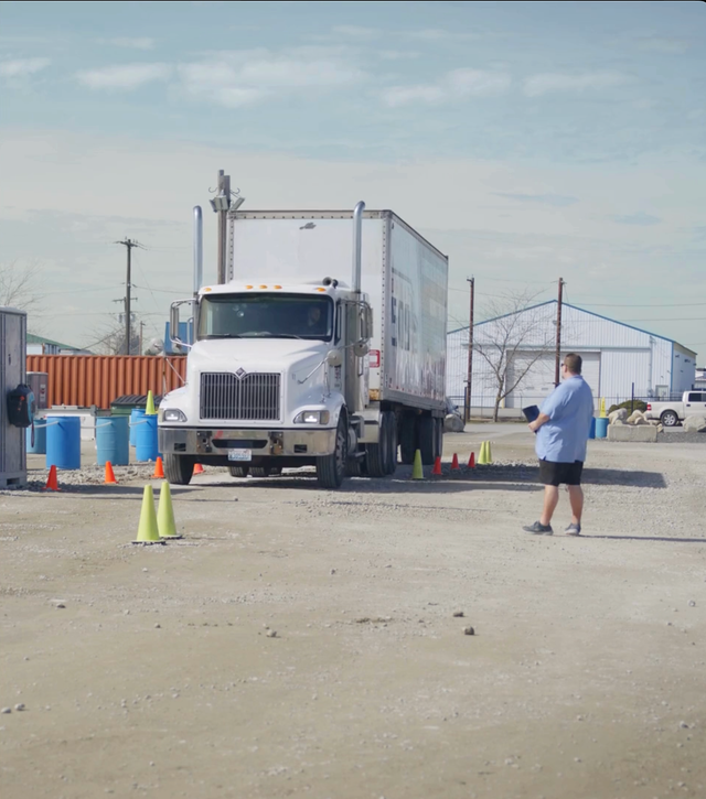 Truck driving training: white semi-truck driving through cones. Instructor watches in a dirt lot on a sunny day.