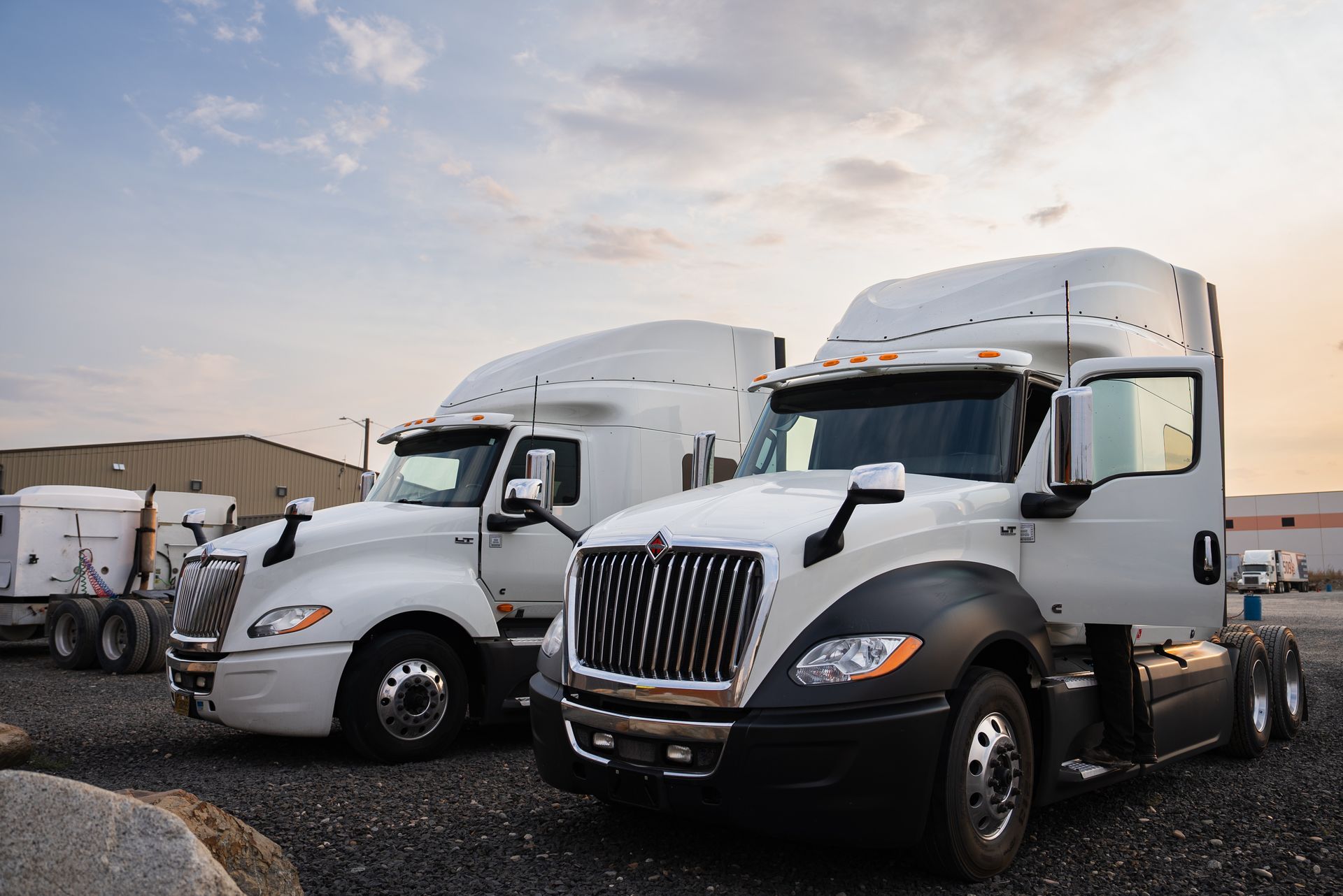 Two white semi-trucks parked outside on gravel at sunset.