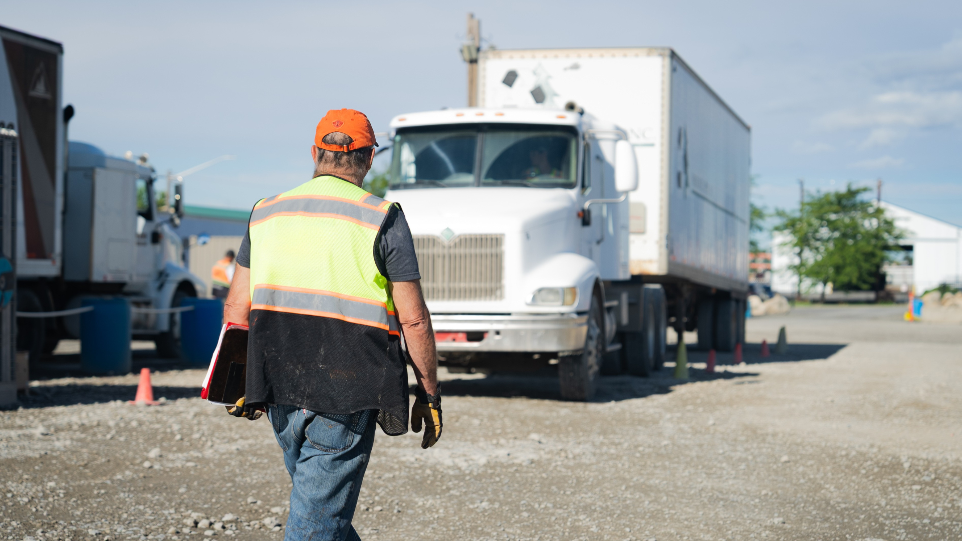 Worker in safety vest walks toward a white semi-truck in a gravel lot on a sunny day.