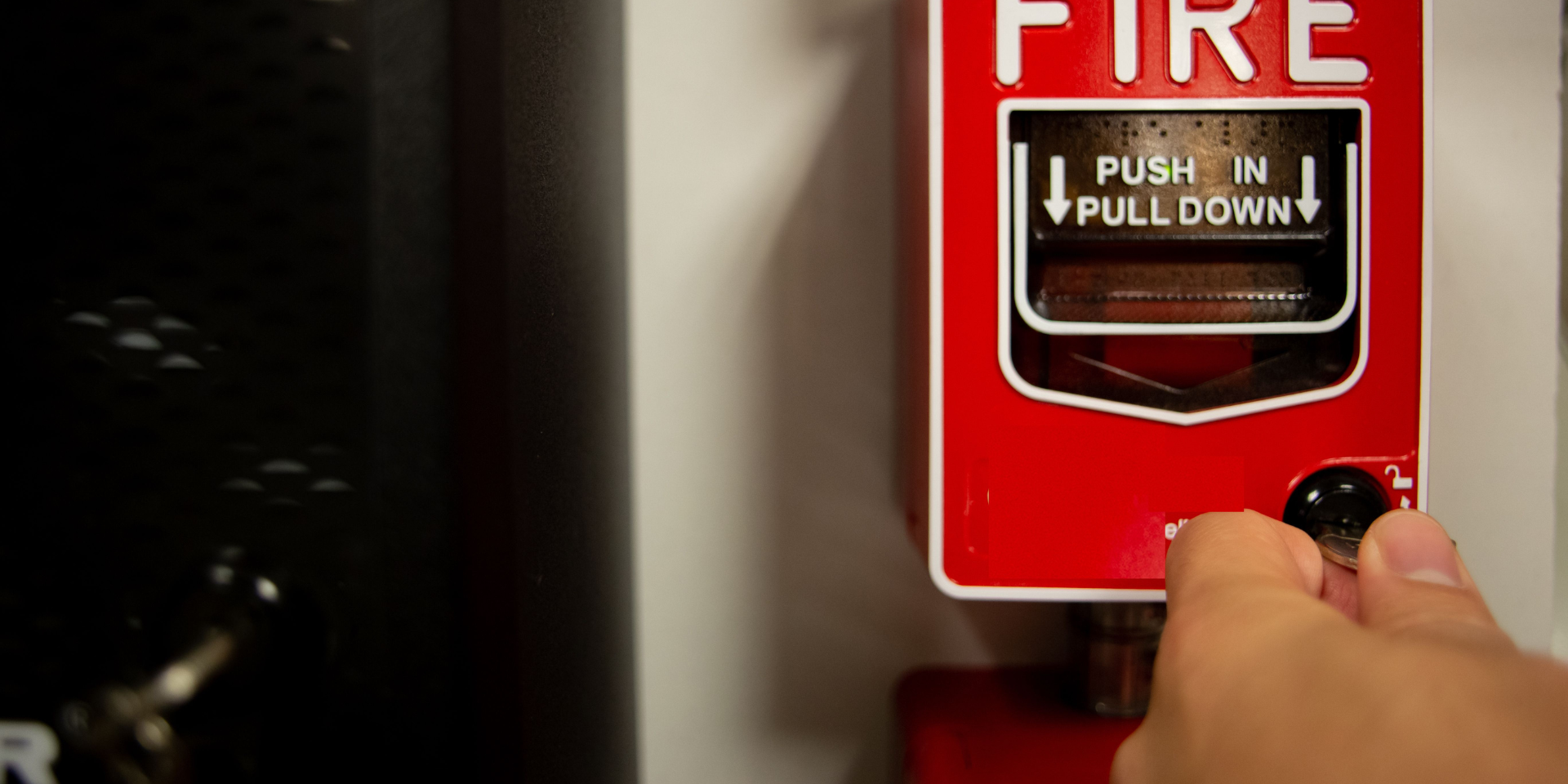 A hand pulling down a fire alarm. Red alarm box on white wall, black object to the left.