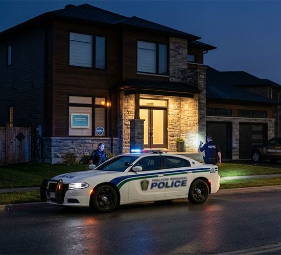 Police car parked outside a lit house at night, with officers near the front door.