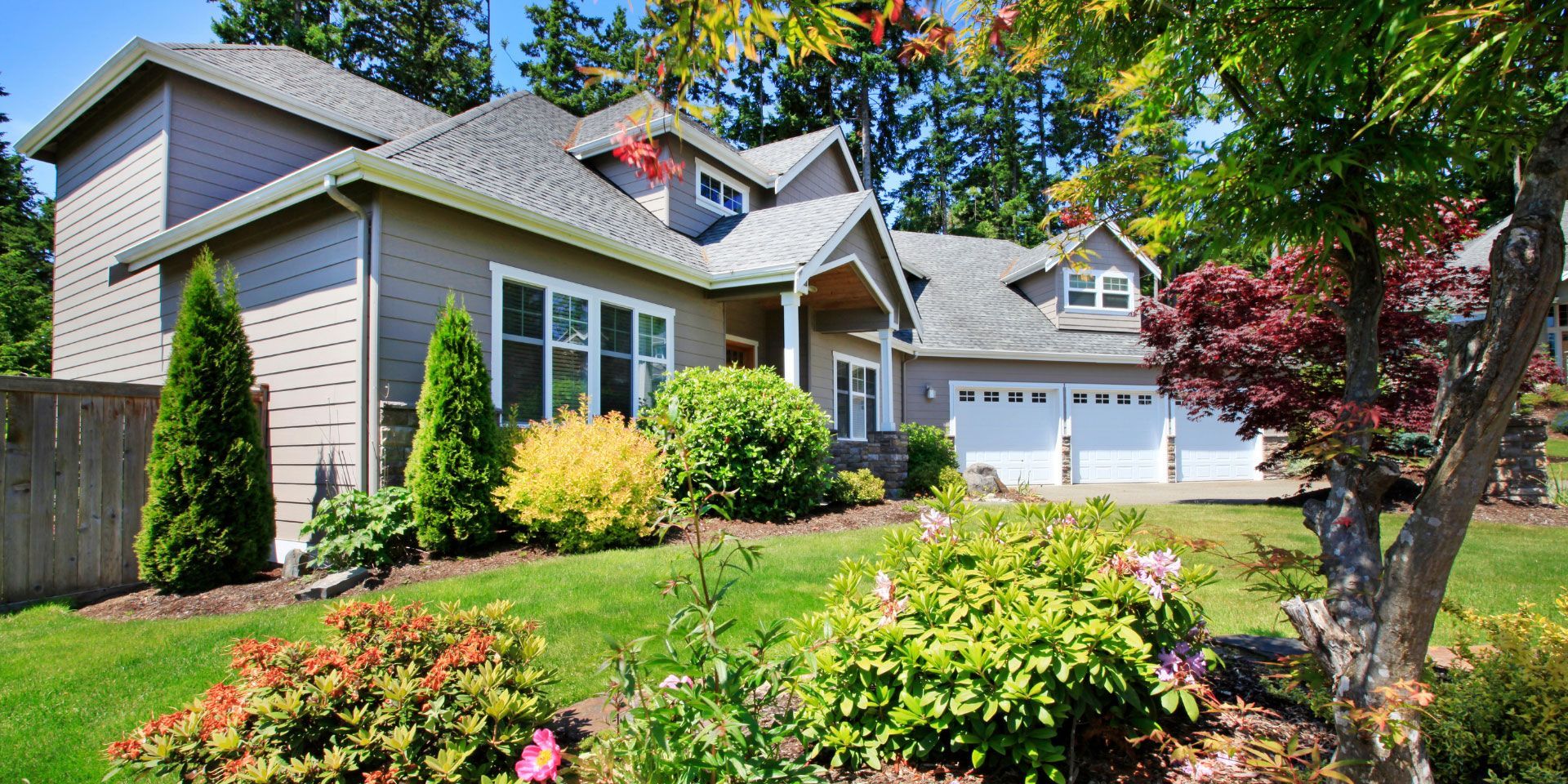 Suburban house with gray siding, white trim, landscaped garden, and driveway under sunny blue sky