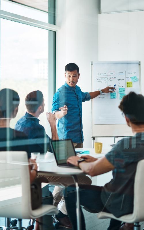 Team meeting in a glass-walled office, presenter pointing at a whiteboard while coworkers listen around a table