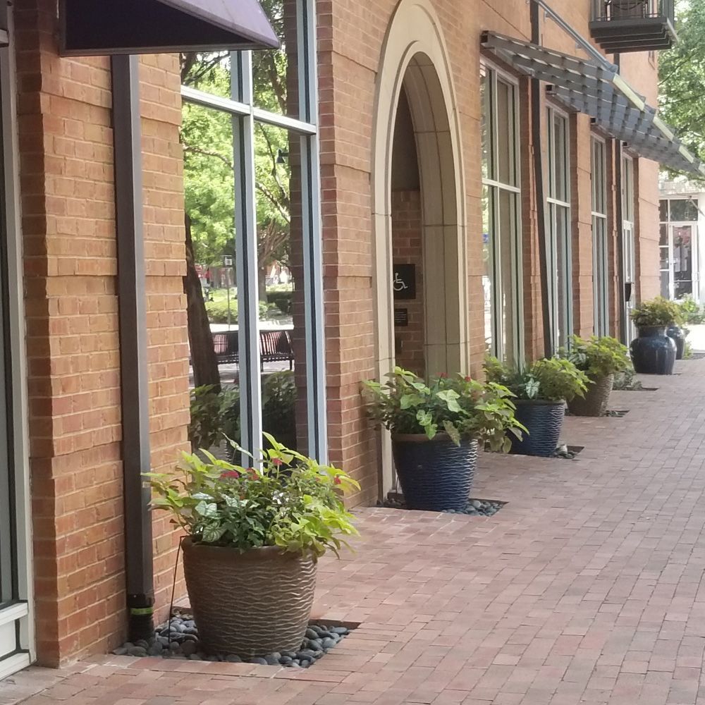 Brick sidewalk beside storefronts with large windows and potted plants along the building.