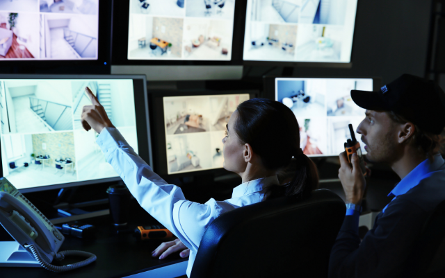 Two security personnel monitoring surveillance footage on multiple screens.