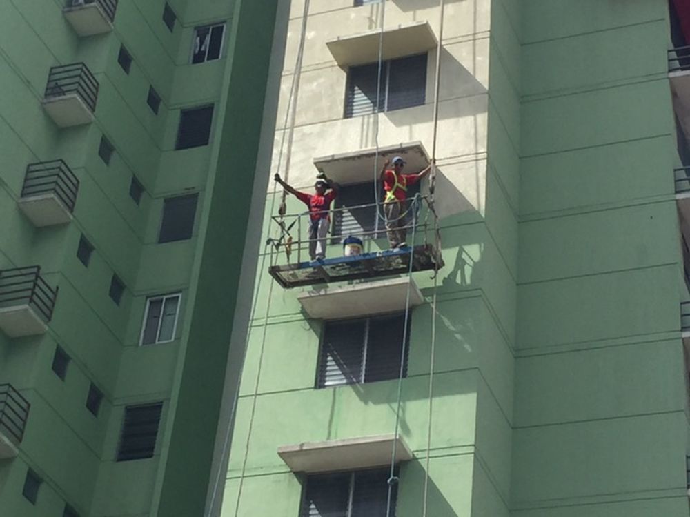 Two workers in red shirts stand on a hanging scaffolding platform against the side of a tall green apartment building.
