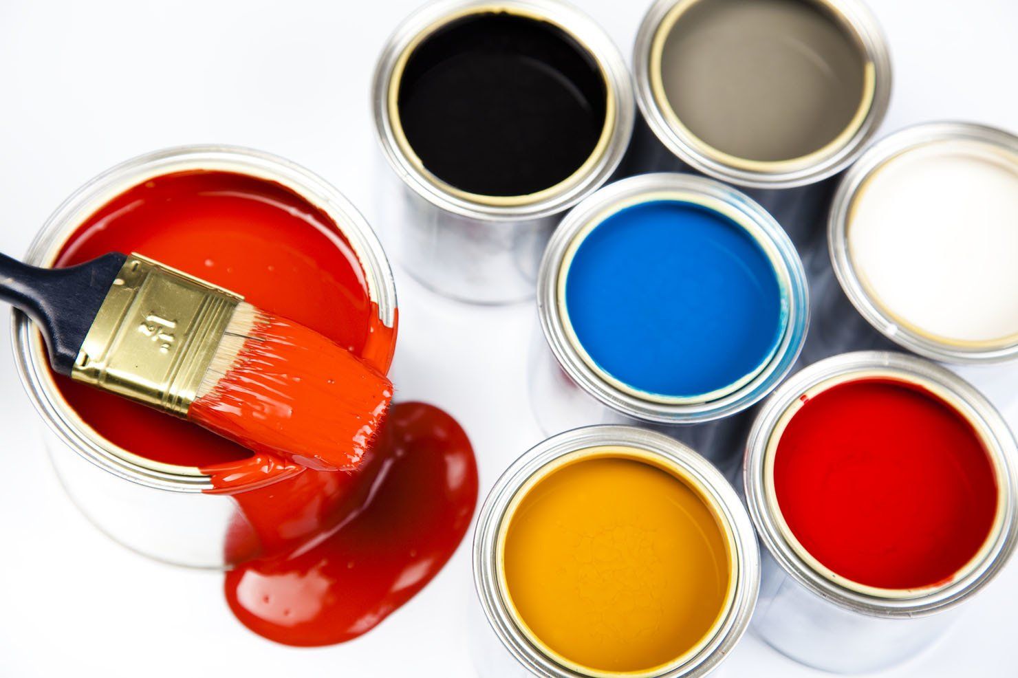 A paintbrush resting in a can of spilled orange paint, surrounded by various other open cans of colored paint.