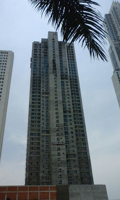 A tall, unfinished concrete high-rise apartment building seen from below, framed by palm fronds against a gray sky.