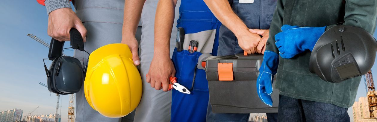 Group of construction workers wearing uniforms, holding safety gear and tools against an outdoor building site background.