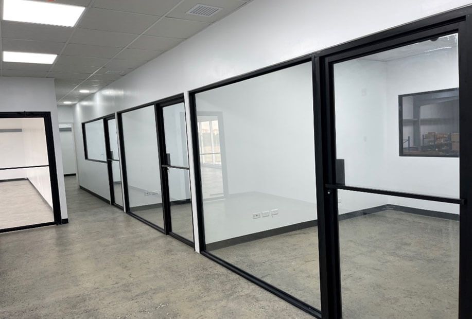 A hallway with concrete floors leads to a series of empty offices enclosed by black-framed glass walls.