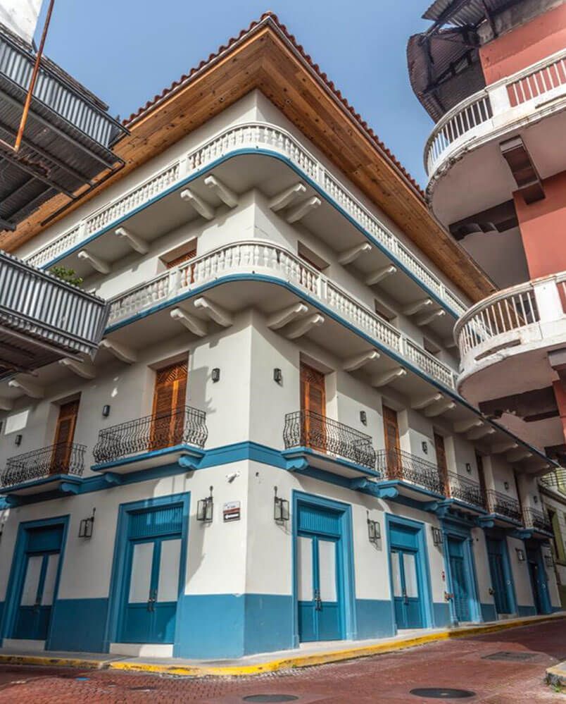 A white, multi-story building with blue trim, wooden doors, and wrap-around balconies on a paved street corner.
