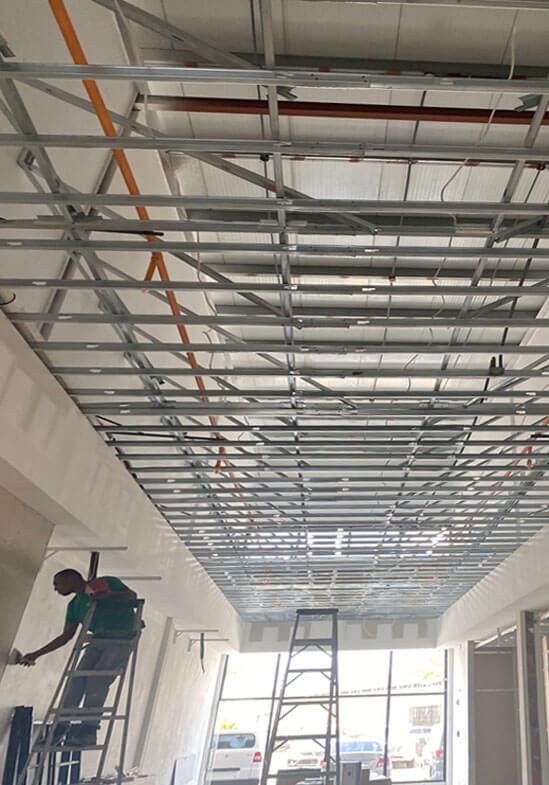 A worker on a stepladder installs metal ceiling grid framing in an unfinished building interior.