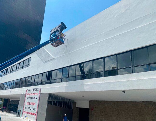 Workers in a lift platform painting a white exterior wall of a building with dark glass windows under a clear blue sky.