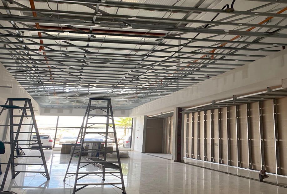 Construction site of an interior space with a suspended metal ceiling grid, ladder equipment, and unfinished wall studs.