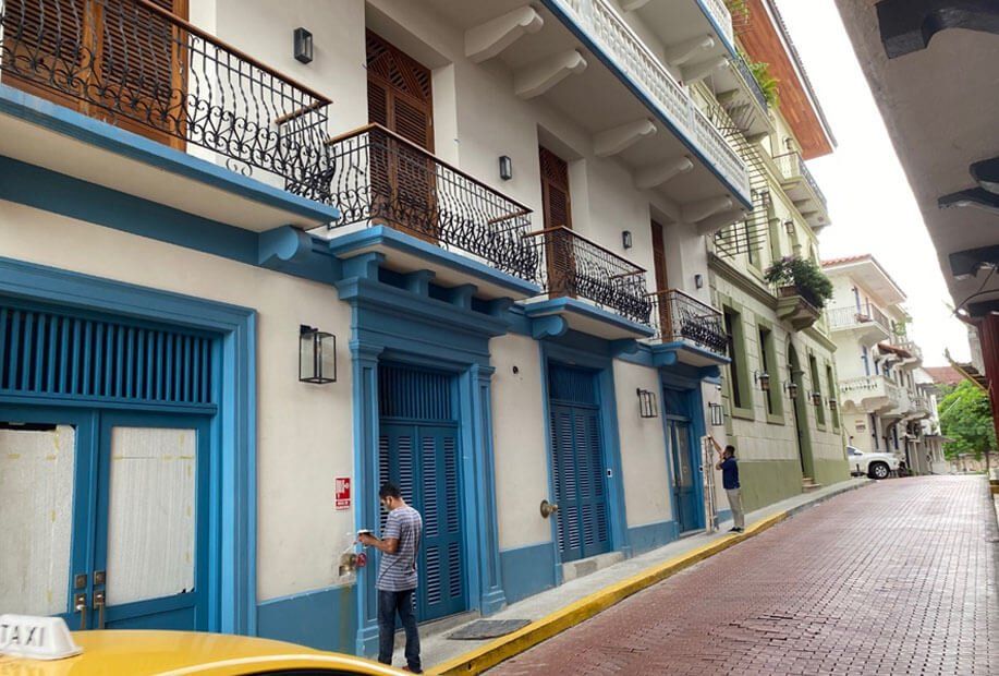 A street view of white colonial buildings with blue trim and iron balconies, with two people standing on a brick road.