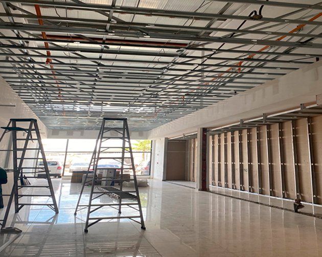 A commercial interior under construction featuring an open metal ceiling grid, ladder scaffolds, and wall framing.