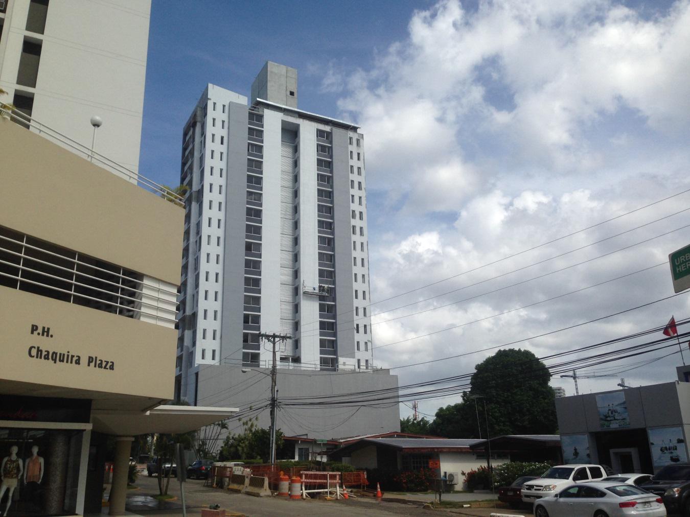 A tall, modern residential tower in white and grey stands behind a tan commercial building under a blue, cloudy sky.