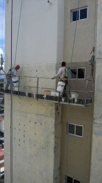 Two workers on a suspended scaffolding platform painting the side of a tall, beige apartment building.