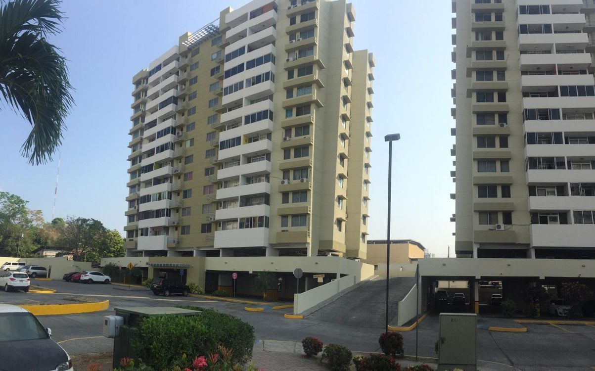 Two tall, light-colored apartment buildings stand side-by-side with parking areas on the ground level.