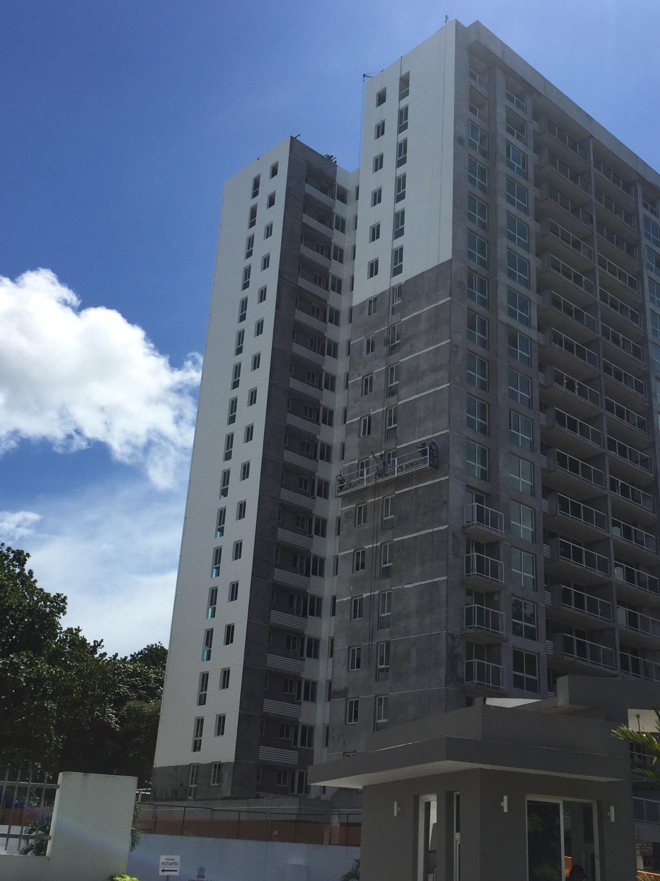 A tall, modern residential apartment building with gray and white concrete exterior walls against a blue, cloudy sky.