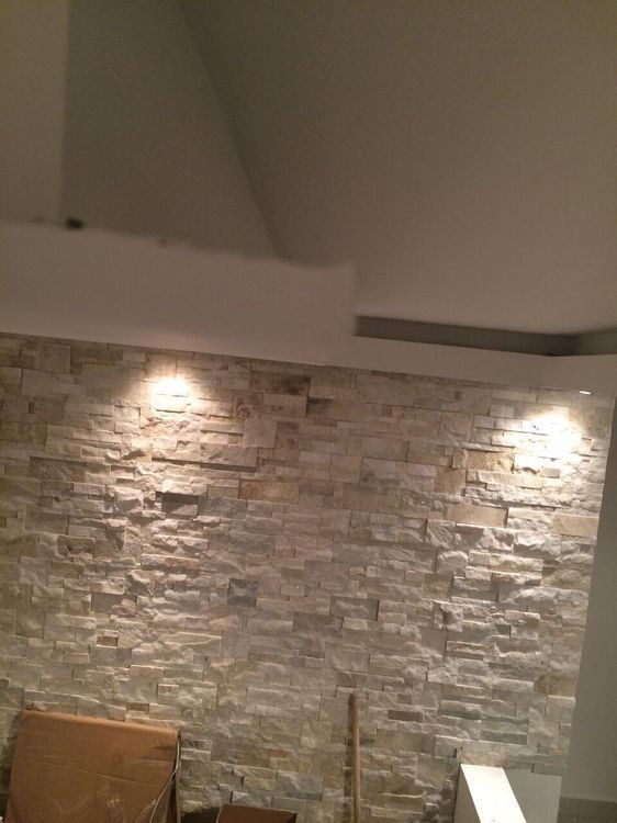 A stone-tiled wall illuminated by two ceiling spotlights in a room with a white angled ceiling.