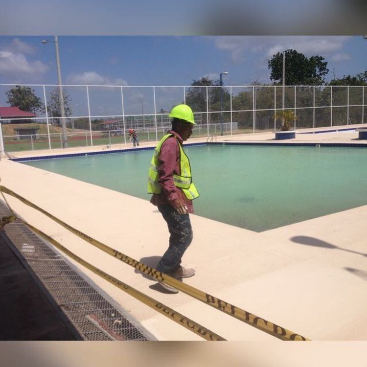 A worker in a high-visibility vest and hard hat walks near a fenced, empty outdoor swimming pool.
