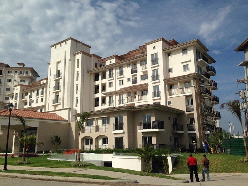 A multi-story beige apartment building with balconies and a tile roof, viewed from a street with two people standing nearby.