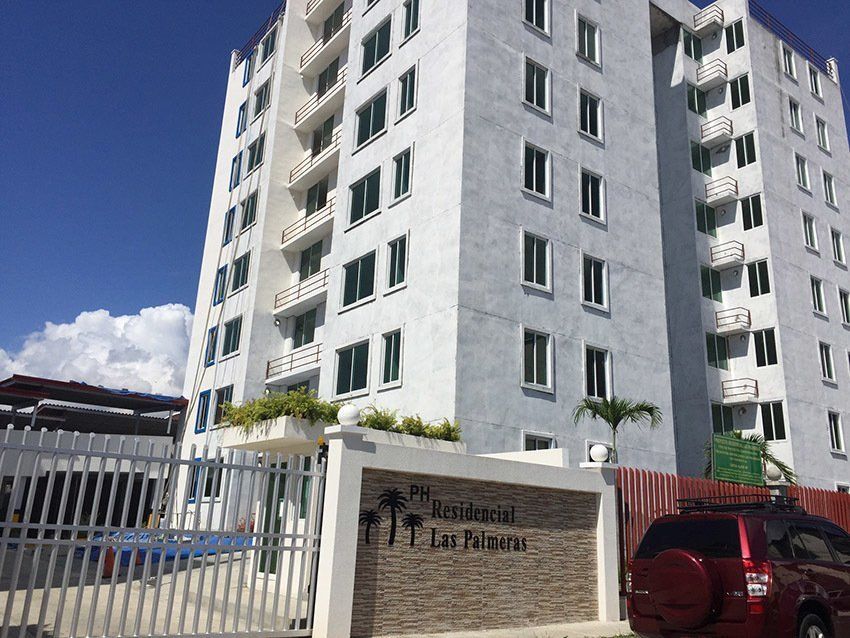 A modern white, multi-story apartment building named PH Residencial Los Palmeras behind a white gate and stone entrance.