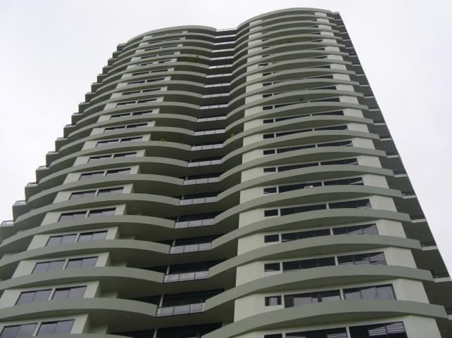 A modern high-rise residential building with a wave-patterned facade and many balconies, viewed from a low angle.