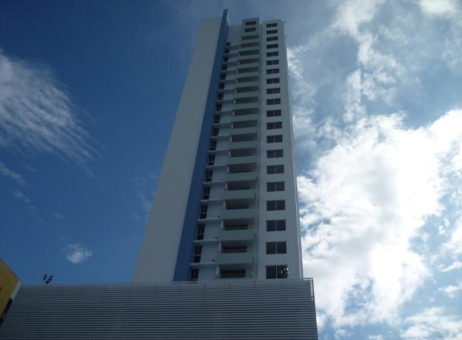 A tall, modern residential skyscraper with white and blue accents, set against a bright, cloudy blue sky.