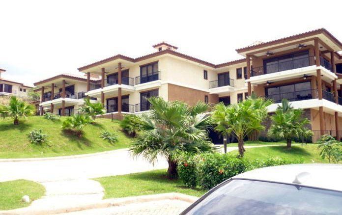 A multi-level apartment complex with beige stucco walls, dark balconies, and a landscaped lawn with palm trees.