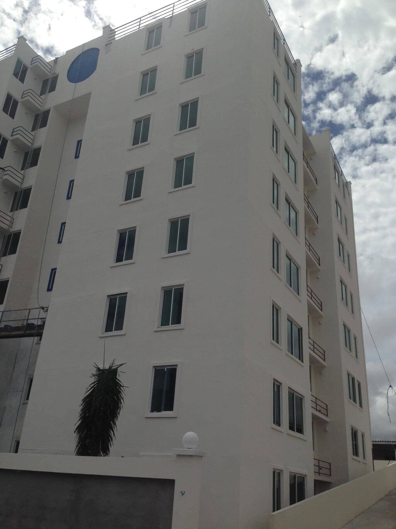 Tall, white modern multi-story apartment building with a blue circular sign, exterior balconies, and cloudy sky.