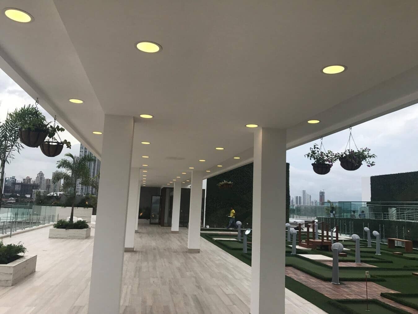 A covered walkway with recessed lighting leads to an outdoor rooftop terrace featuring potted plants and a putting green.