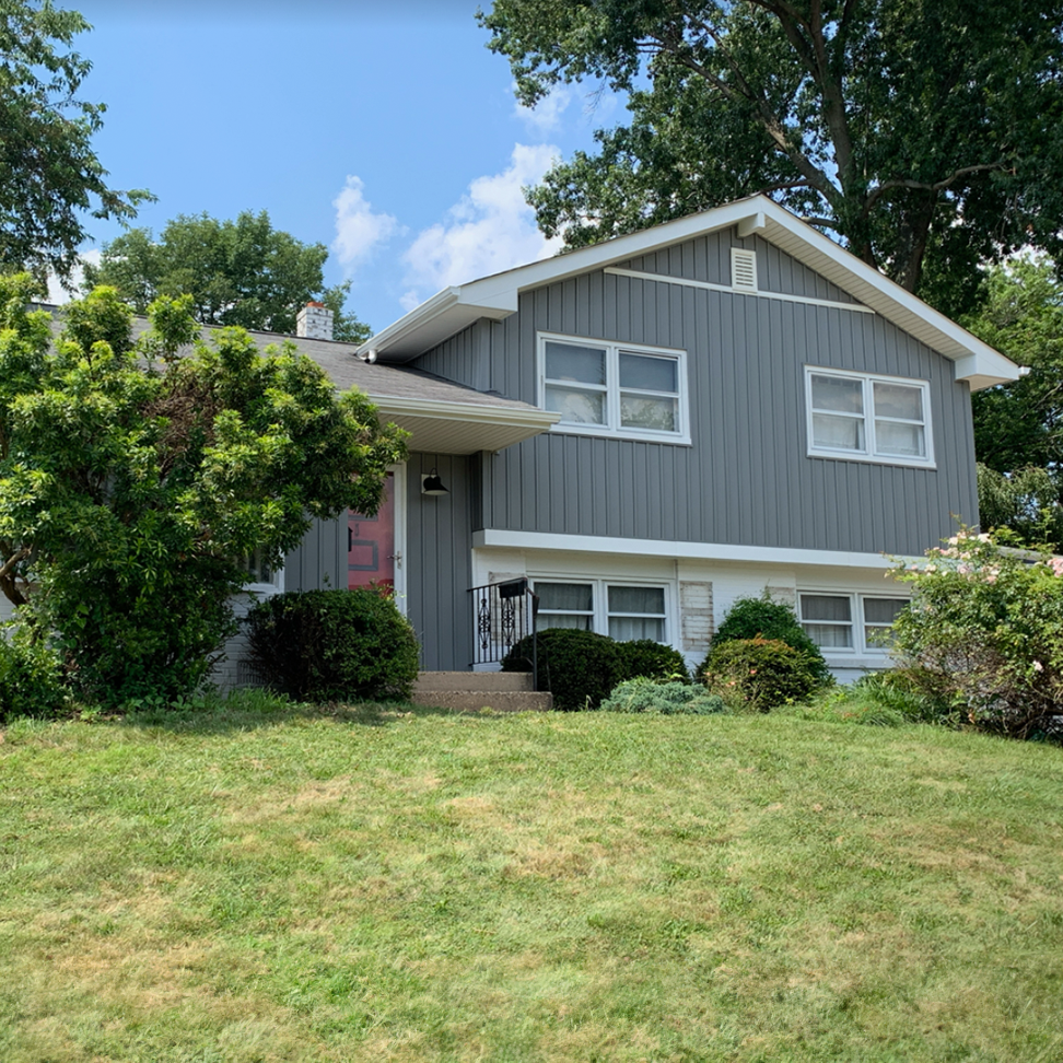 a nicely updated gray house with white trim sits on a lush green hillside with nice curb appeal!