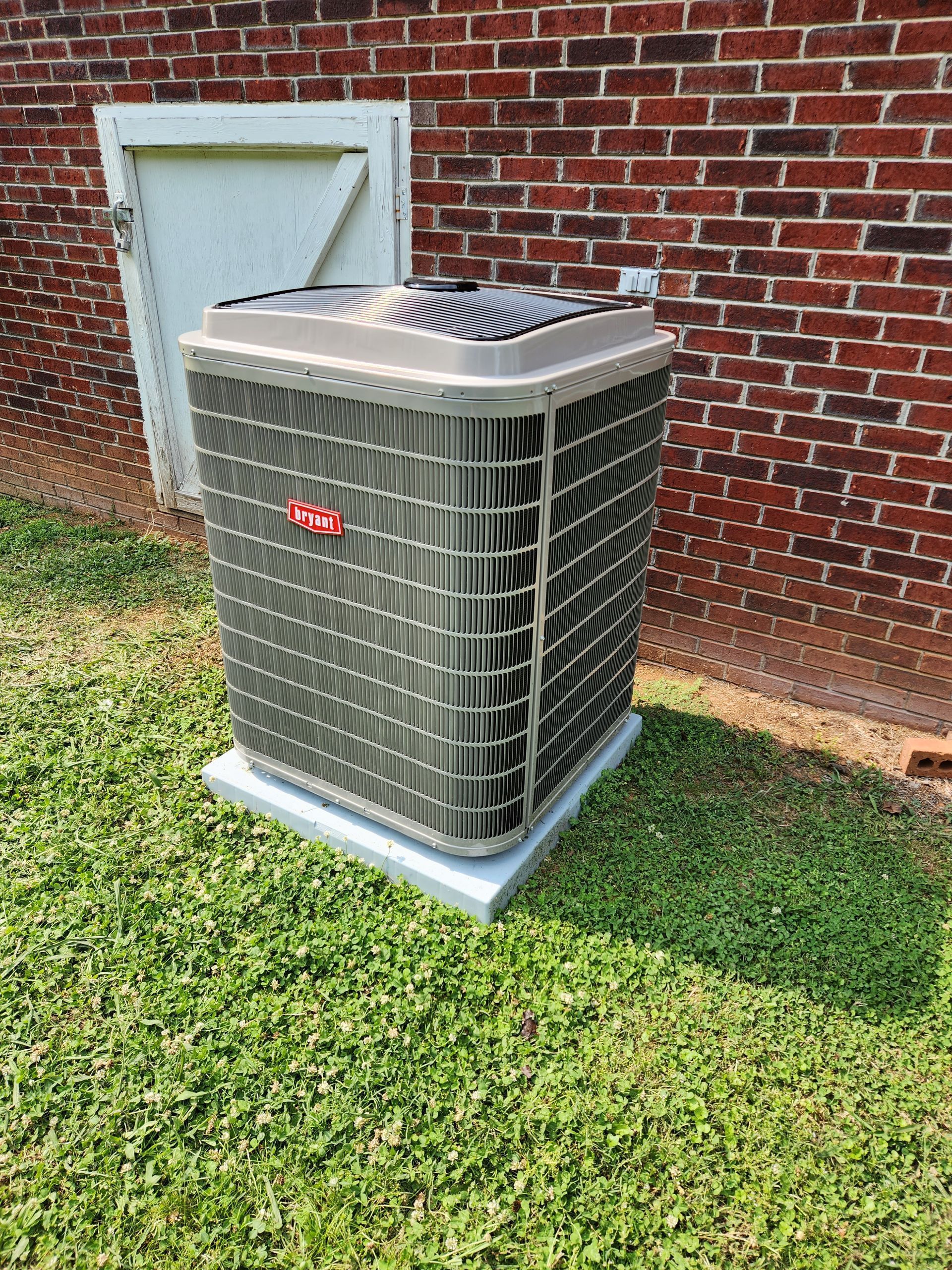 Outdoor air conditioner unit on grass beside a brick wall and utility door.