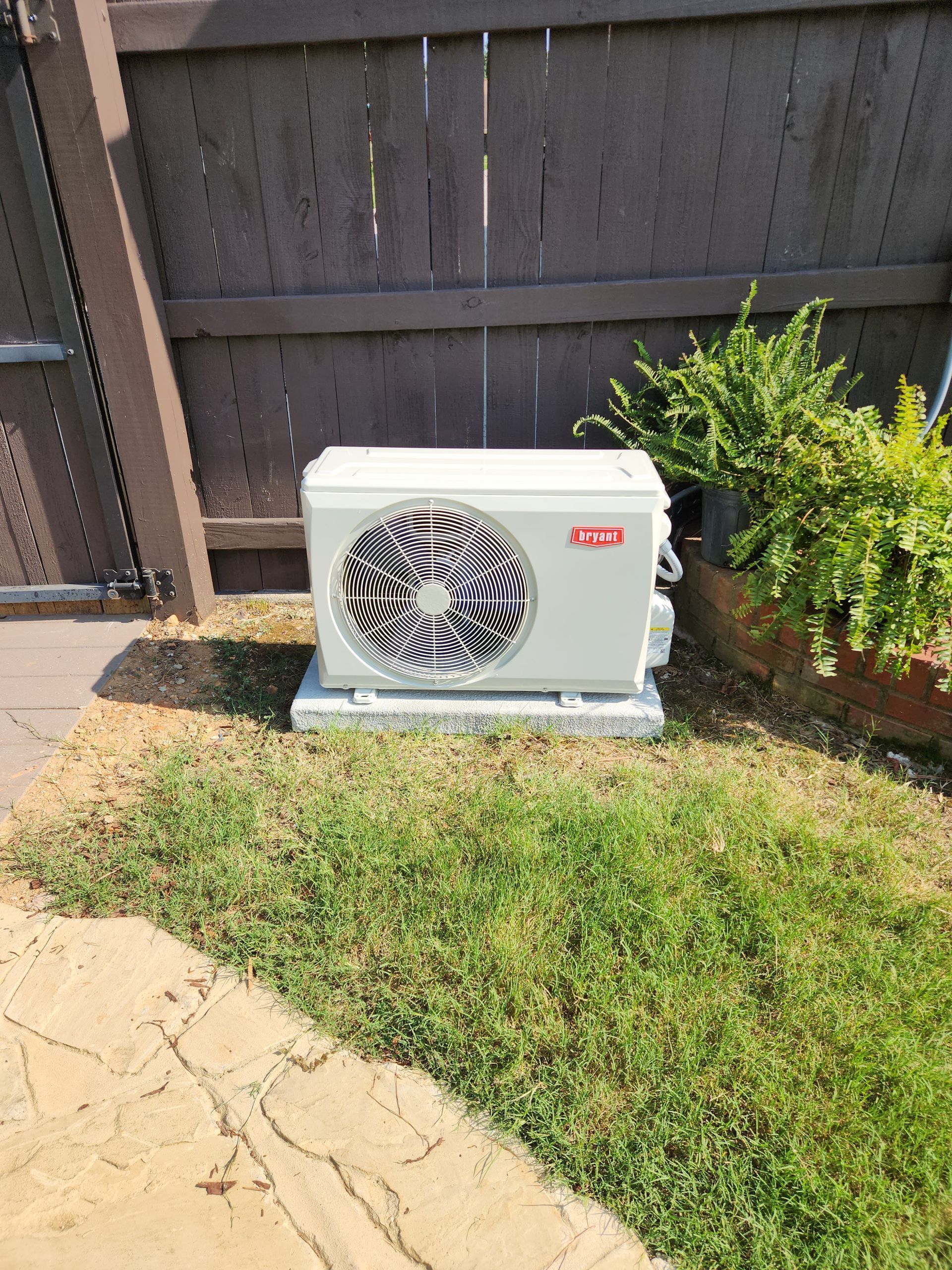 Outdoor HVAC unit beside a wooden fence and green shrub on a sunny patio.