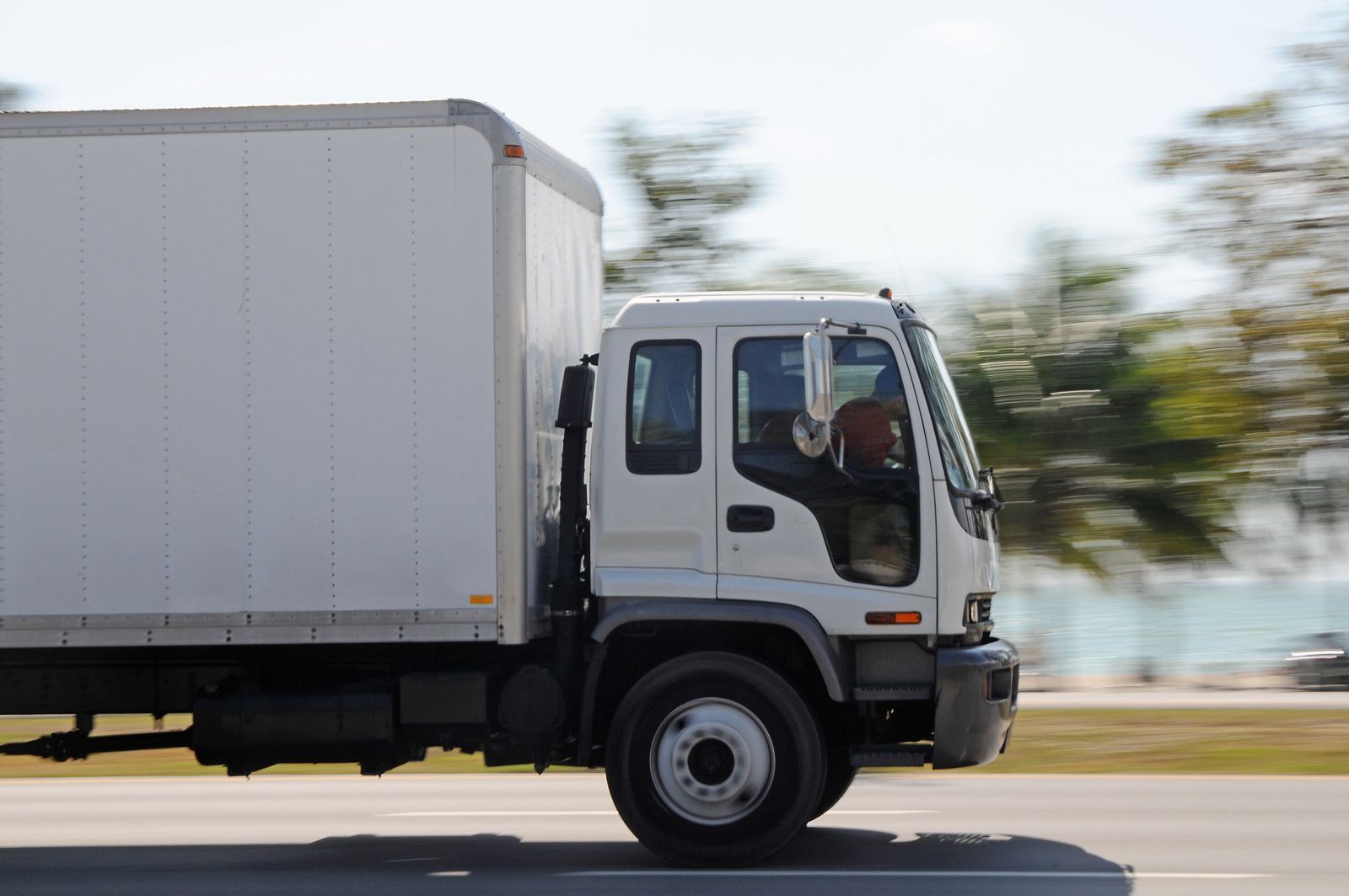 White box truck driving on a road, trees blurred in the background.