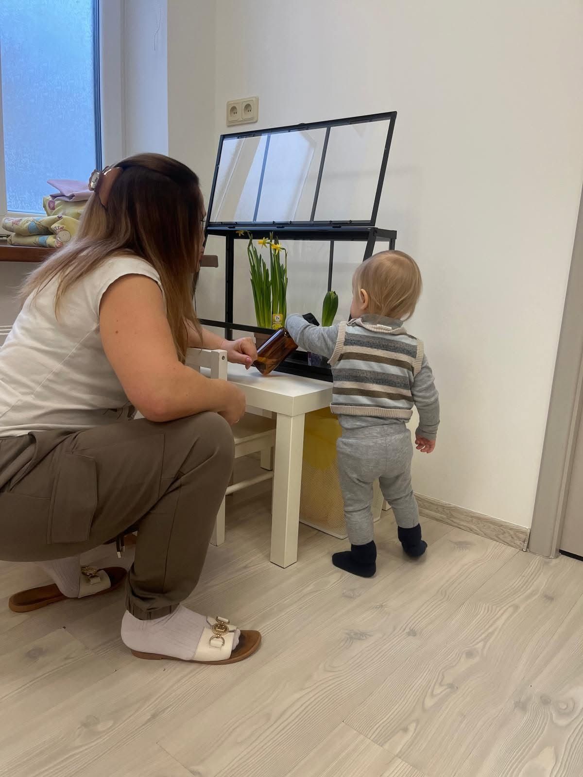Pieaugušais pietupies pie galdiņa pie loga līdzās mazulim, abi aplūko augus un papīrus.