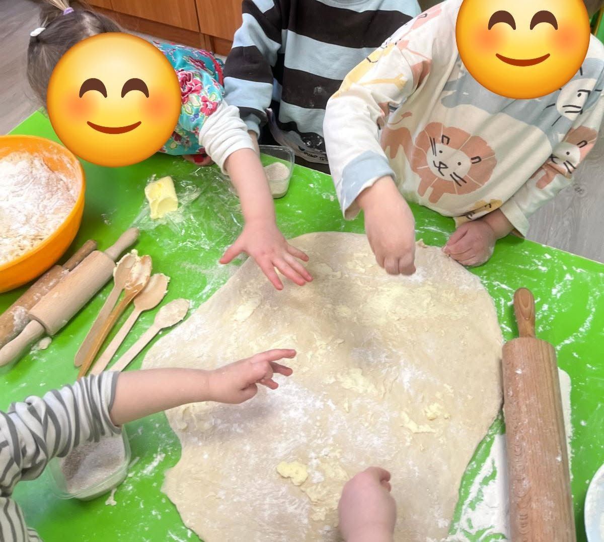 Bērni mīca mīklu pie galda, kas nokaisīts ar miltiem, izmantojot mīklas rullīšus un koka karotes.
