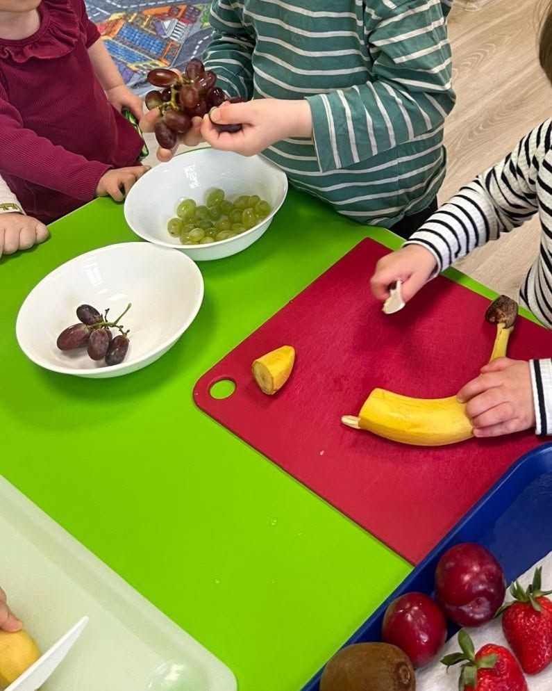 Bērni klasē gatavo augļus uz krāsainiem galdiem, piedaloties uzkodu gatavošanas aktivitātē