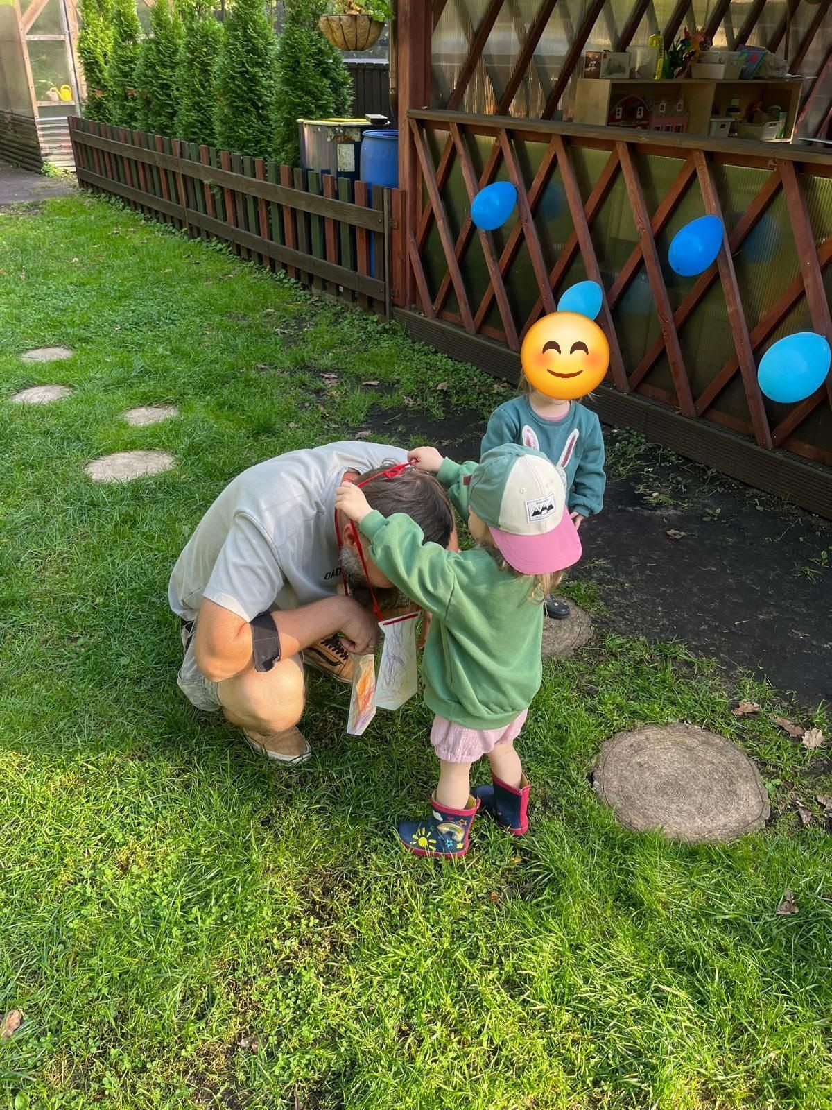 Divi bērni spēlējas ārā uz zāles pie koka žoga — viens ir pietupies, bet otrs sniedzas viņa virzienā.