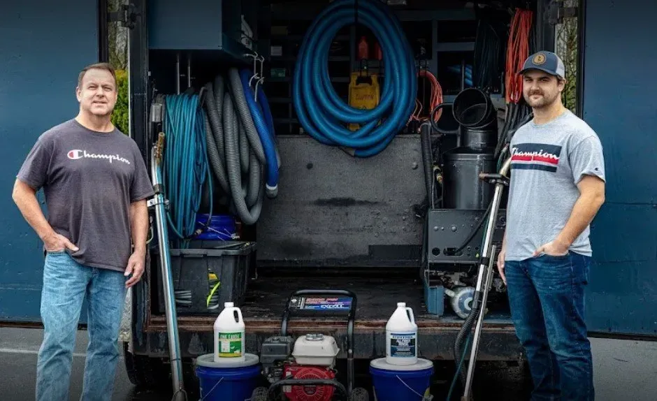 Two men standing beside a carpet-cleaning truck with hoses and equipment