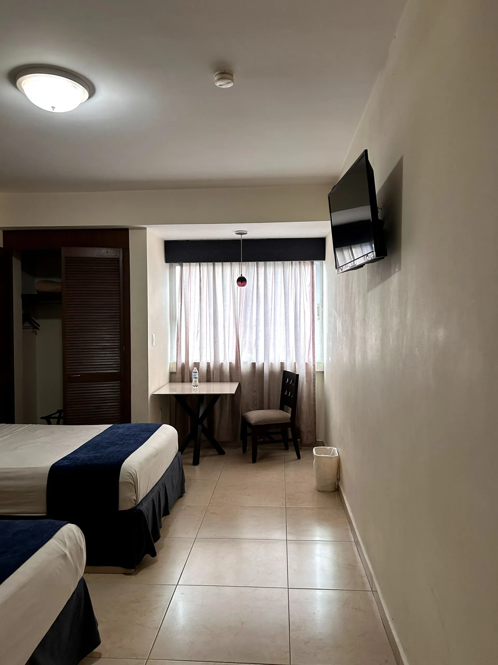 Habitación de hotel con dos camas, una mesita con silla junto a una ventana con cortinas y un televisor montado en la pared.