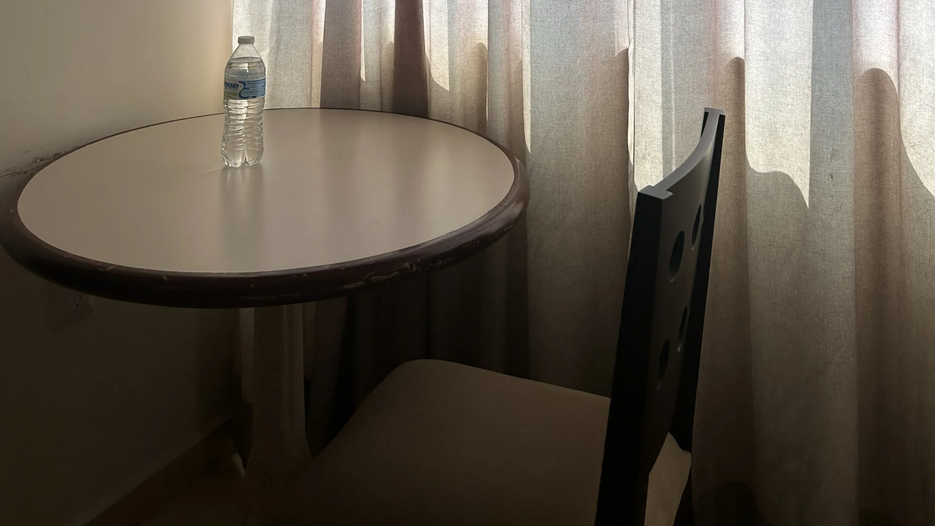Una pequeña mesa redonda con una botella de agua se encuentra junto a una silla de madera oscura, frente a unas cortinas.