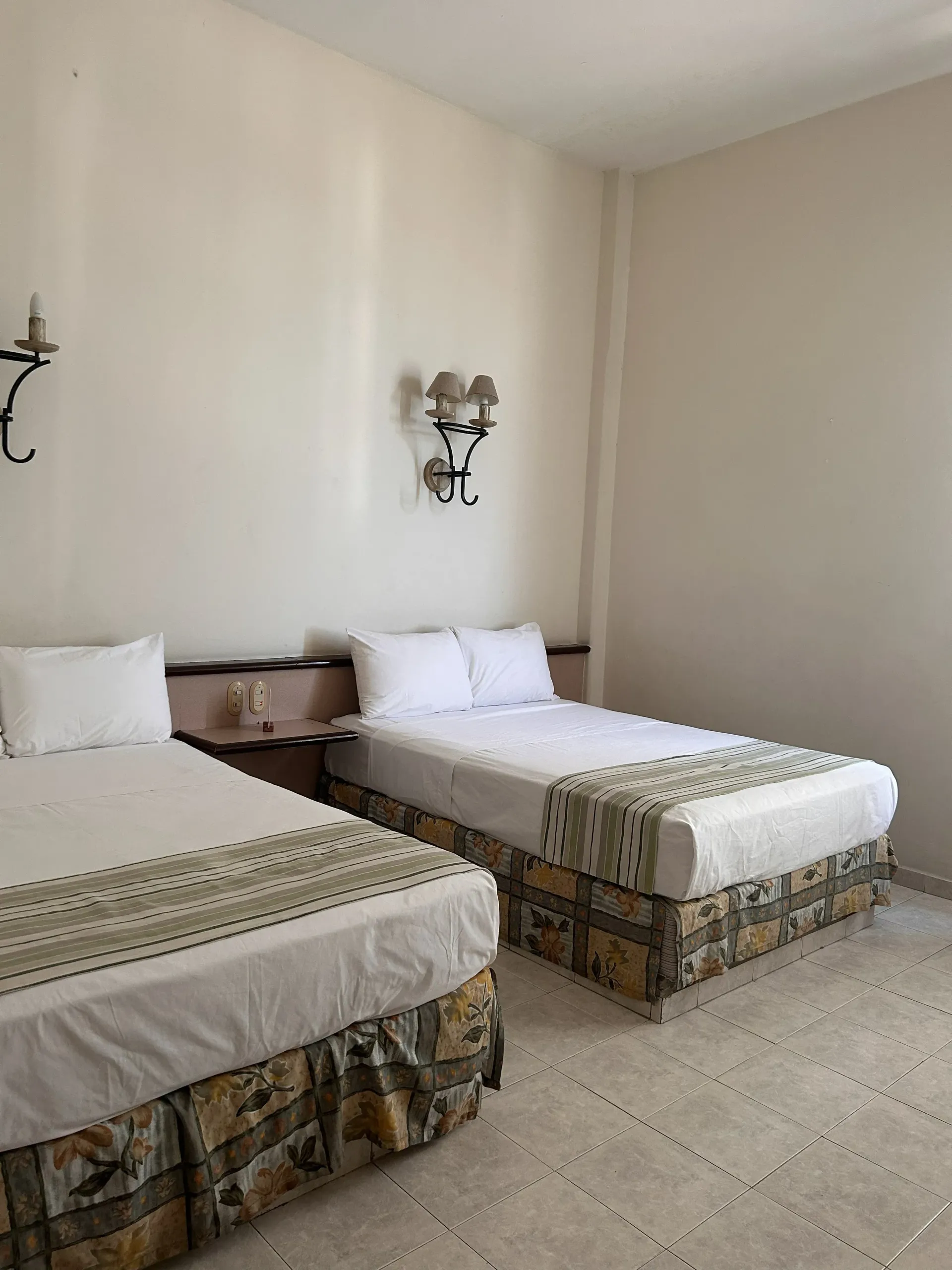 Dos camas de hotel con sábanas blancas y faldones de cama marrones estampados en una habitación beige con apliques de pared.